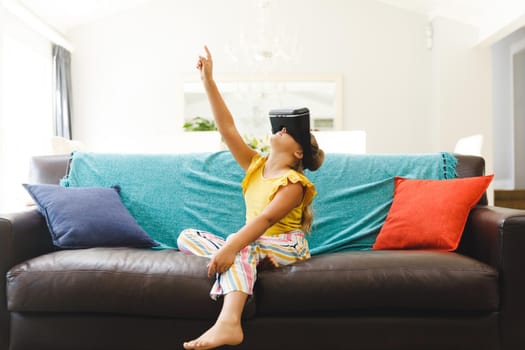 Caucasian girl sitting on couch and using vr headset in living room. childhood leisure time, fun and discovery at at home using technology.