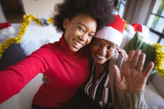 Smiling african american adult daughter and senior mother in santa hats taking christmas selfie. christmas, festivity and communication technology at home.