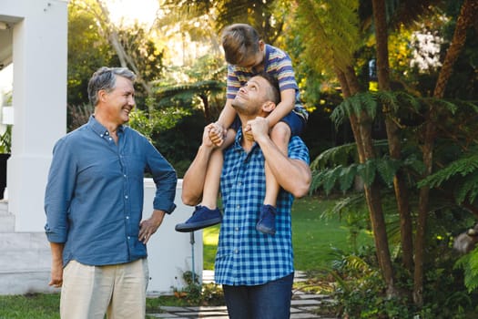 Portrait of smiling caucasian grandfather and adult son with grandson on his shoulders in garden. multi generation family enjoying leisure time together at home.