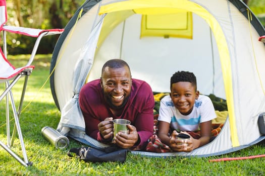 Portrait of african american father with son having fun and lying in tent with drinks in garden. family spending time at home.