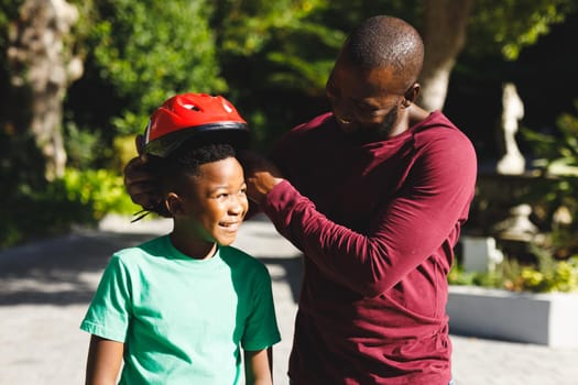 African american father with son smiling and putting on helmet before skateboarding in garden. family spending time at home.