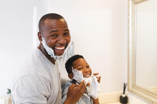 Portrait of smiling african american father with son having fun with shaving foam in bathroom. family spending time at home.
