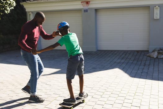 African american father smiling and helping son balancing on skateboard in garden. family spending time at home.