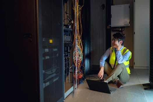Asian male engineer wearing high visibility vest with laptop inspecting in computer server room. database server management and maintenance concept