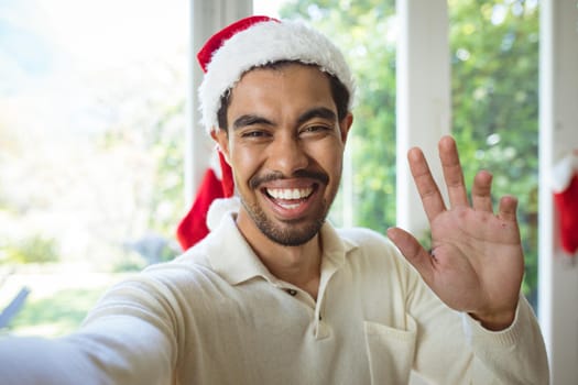 Portrait of happy biracial man in santa hat making christmas video call. christmas, festivity and communication technology.