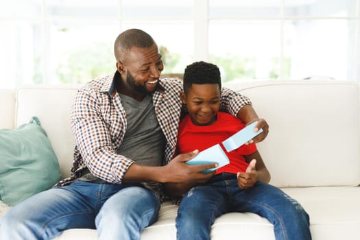 Happy african american father opening gift from his son and smiling in living room. family spending time at home, father son relationship.