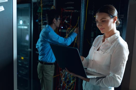 Caucasian female engineer using laptop and asian male engineer inspecting in computer server room. database server management and maintenance concept