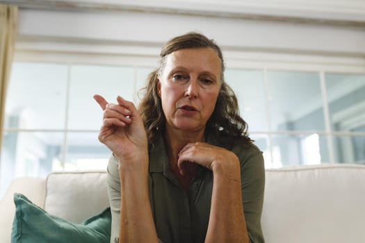 Senior caucasian woman in living room, sitting on sofa, talking and gesturing during video call. retirement lifestyle, spending time at home with technology.