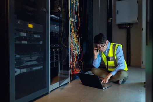 Asian male engineer using laptop and talking on smartphone while inspecting in computer server room. database server management and maintenance concept