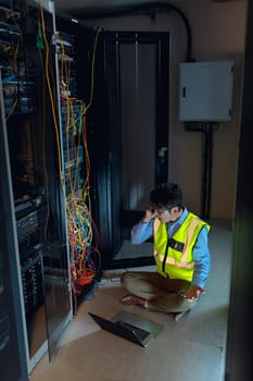 Asian male engineer with laptop talking on smartphone while inspecting in computer server room. database server management and maintenance concept