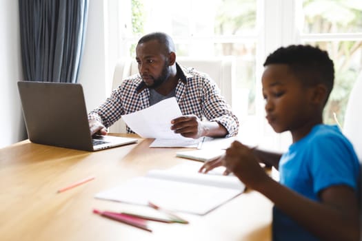 African american father working on laptop in dining room with son sitting with him doing homework. family spending time at home.