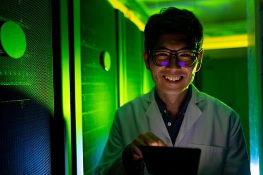 Portrait of asian male engineer wearing an apron with digital tablet smiling in computer server room. database server management and maintenance concept