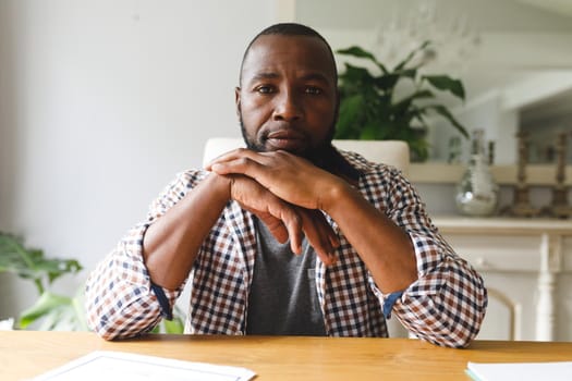 Serious african american man sitting at table in dining room, listening during video call. flexible working from home with technology.