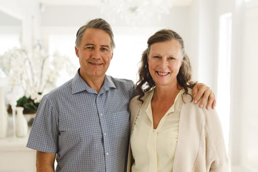 Portrait of happy senior caucasian couple in living room, embracing and smiling. retirement lifestyle, spending time at home.