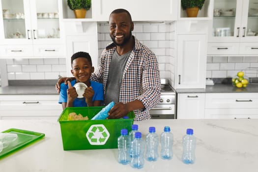 Portrait of happy african american father and son in kitchen sorting rubbish for recycling. family spending time at home.