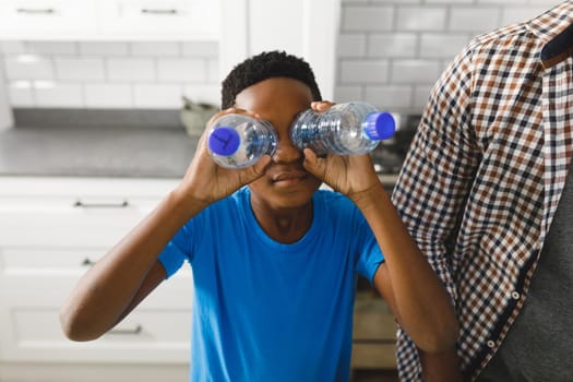 Happy african american son sorting recycling with father in kitchen, playing with plastic bottles. family spending time at home.