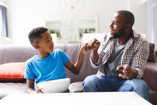 Happy african american father and son sitting on sofa, fist bumping watching tv. family spending time together at home.