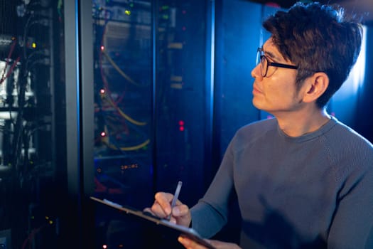 Asian male engineer writing on clipboard while inspecting in computer server room. database server management and maintenance concept
