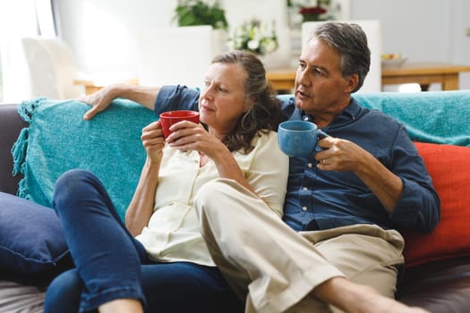 Happy senior caucasian couple in living room sitting on sofa, drinking coffee. retirement lifestyle, spending time at home.