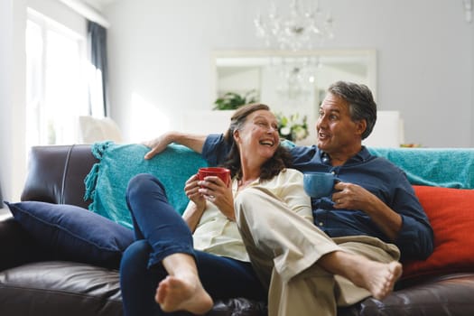 Happy senior caucasian couple in living room sitting on sofa, drinking coffee. retirement lifestyle, spending time at home.