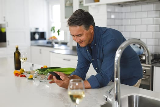 Smiling senior caucasian man in modern kitchen, using smartphone. retirement lifestyle, spending time at home.