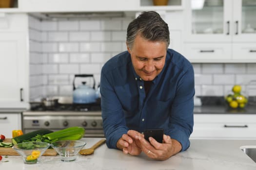 Smiling senior caucasian man in modern kitchen, using smartphone. retirement lifestyle, spending time at home.