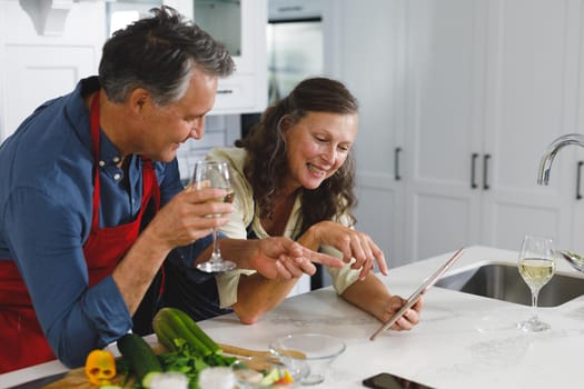 Happy senior caucasian couple in kitchen cooking together, drinking wine, using tablet. retirement lifestyle, spending time at home.