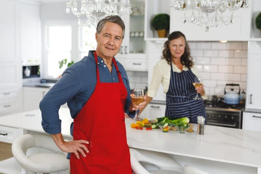 Portrait of happy senior caucasian couple in kitchen cooking together, drinking wine. retirement lifestyle, spending time at home.