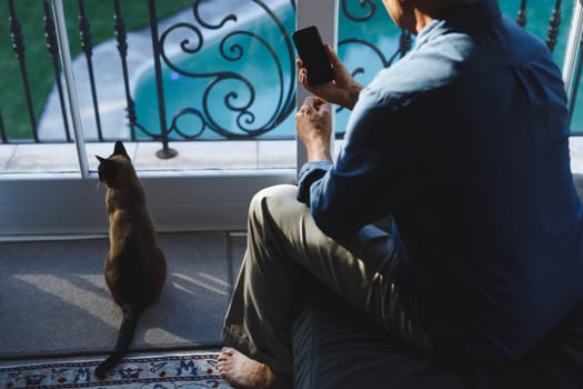 Senior caucasian man with cat in living room, sitting on sofa, using smartphone. retirement lifestyle, spending time at home with technology.