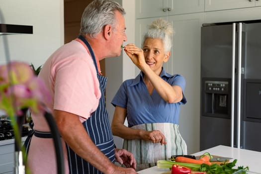 Happy caucasian senior couple standing in kitchen, preparing meal together and eating vegetables. healthy retirement lifestyle at home.