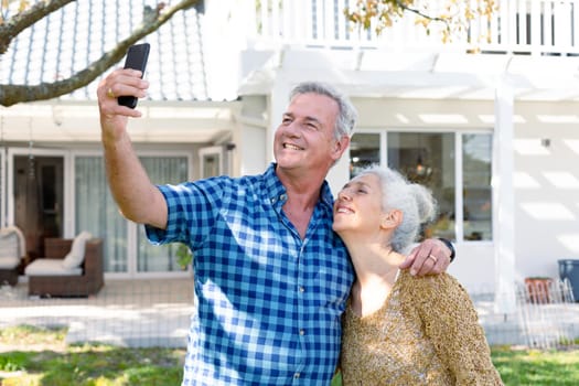 Happy caucasian senior couple taking selfie in front of house. healthy retirement lifestyle at home and garden.
