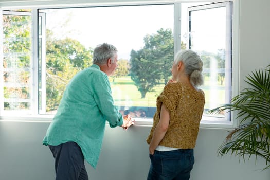 Back view of caucasian senior couple standing at window and talking. healthy retirement lifestyle at home.