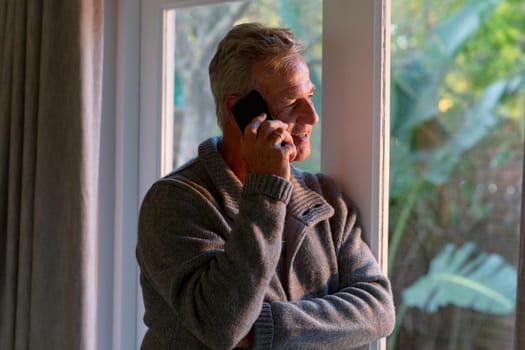 Happy caucasian senior man standing at window and making call. active retirement lifestyle at home.