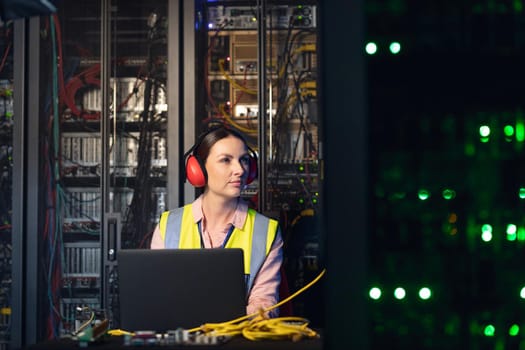 Caucasian female engineer wearing ear plugs using a laptop in computer server room. database server management and maintenance concept