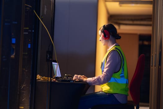 Caucasian female engineer wearing ear plugs using a laptop in computer server room. database server management and maintenance concept