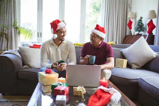 Happy biracial adult son and senior father in santa hats making laptop christmas video call. christmas, festivity and communication technology.