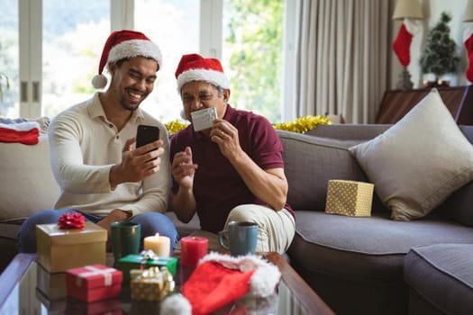 Happy biracial adult son and senior father in santa hats making smartphone christmas video call. christmas, festivity and communication technology.