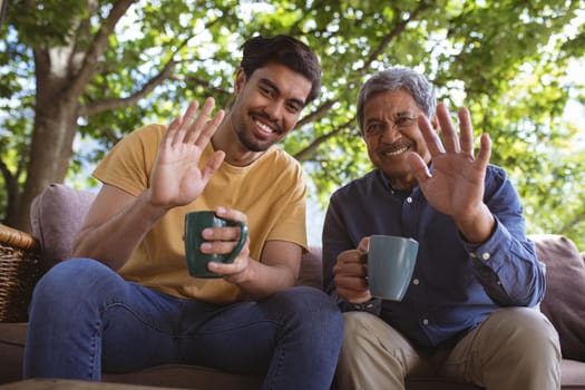 Happy biracial adult son and senior father holding cups of coffee and making laptop video call. family time at home using technology together.