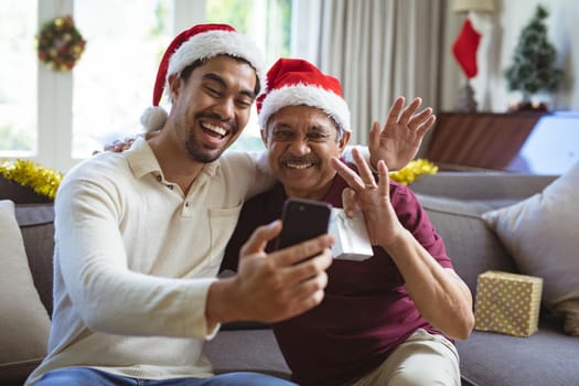 Happy biracial adult son and senior father in santa hats making smartphone christmas video call. christmas, festivity and communication technology.