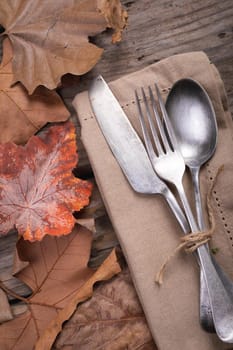 Close up of multiple autumn leaves and cutlery set over a napkin on wooden surface. autumn season and restaurant concept