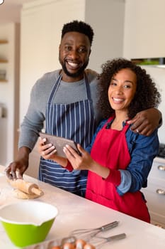 Happy african american couple wearing aprons, baking together and using tablet. family time, having fun together at home.