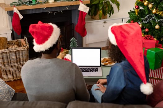 Back view of african american couple wearing santa hats, using laptop with copy space on screen. christmas, festivity and communication technology.