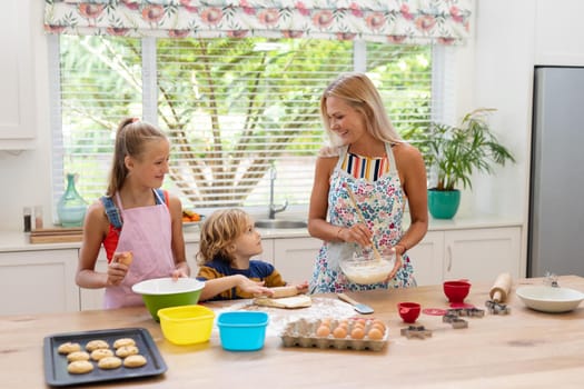 Happy caucasian mother in kitchen with daughter and son, wearing aprons baking cookies together. staying at home in isolation during quarantine lockdown.