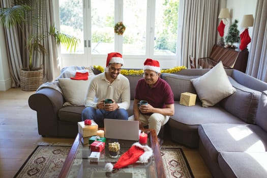 Happy biracial adult son and senior father in santa hats making laptop christmas video call. christmas, festivity and communication technology.