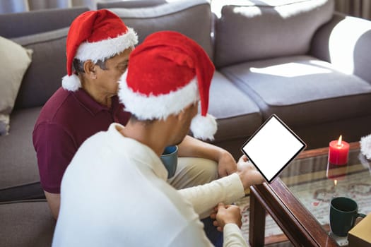 Biracial adult son and senior father in santa hats making tablet christmas video call. christmas, festivity and communication technology.