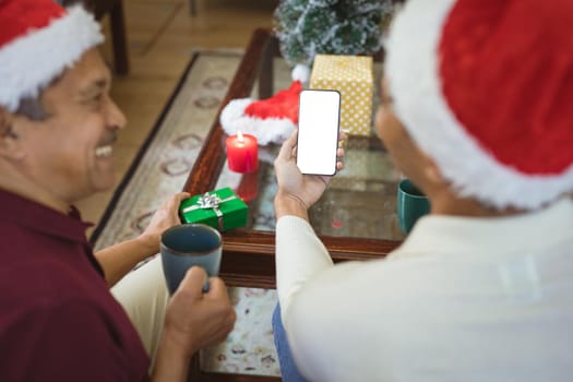 Happy biracial adult son and senior father in santa hats making smartphone christmas video call. christmas, festivity and communication technology.