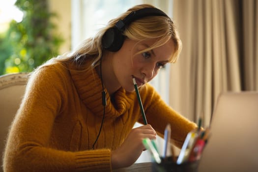 Caucasian woman working in living room at home, wearing headphones and using laptop, concentrating. flexible working from home with technology.