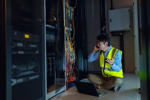 Asian male engineer with laptop talking on smartphone while inspecting in computer server room. database server management and maintenance concept