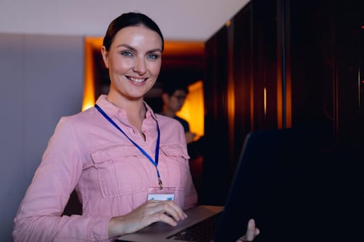 Portrait of caucasian female engineer smiling while using laptop in computer server room. database server management and maintenance concept