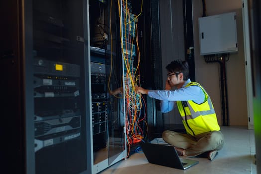 Asian male engineer with laptop talking on smartphone while inspecting in computer server room. database server management and maintenance concept